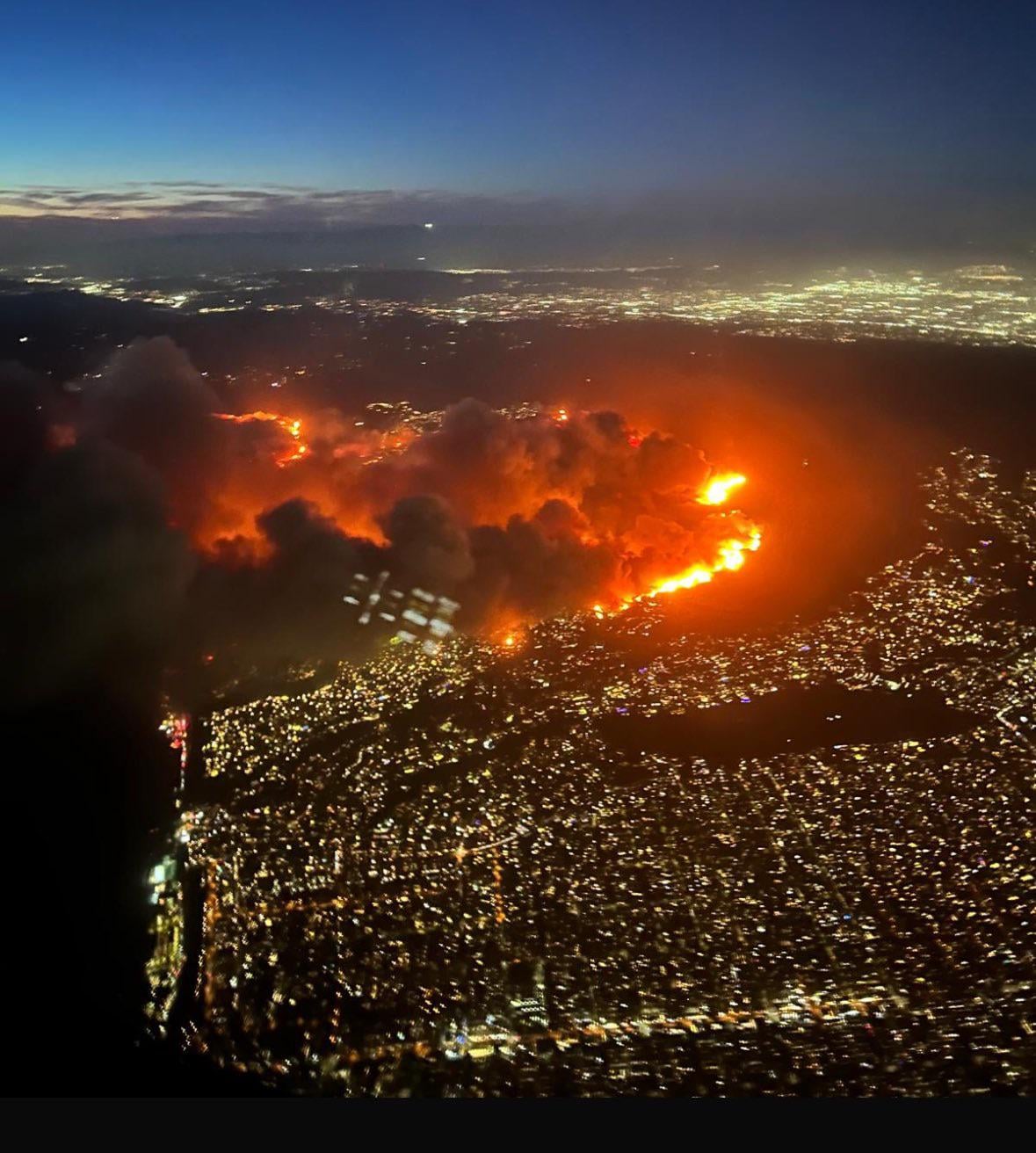 Aerial view of a wildfire burning through neighborhoods above a lit city at dusk